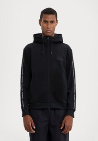 Sudadera con cremallera negra y capucha, con un logo texturizado en el pecho y una marca impresa a lo largo de las mangas. Tela suave, corte relajado.