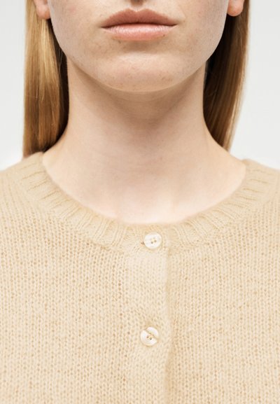 Γυναίκα με ίσια ξανθά μαλλιά που φοράει μια πλεκτή ζακέτα σε κρεμ απόχρωση με δύο ορατά μαργαριταρένια κουμπιά κλειστά στο λαιμό.