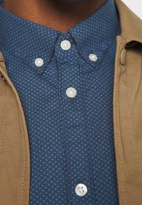 Giacca esterna marrone sopra una camicia blu scuro con motivo e colletto button-down. Presenta bottoni di plastica bianchi e un design geometrico sottile.