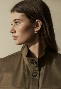 Khaki skjorte med krave og knaplukning foran, der har to frontlommer og en glat tekstur. Modellen har mellemlangt hår og små øreringe.