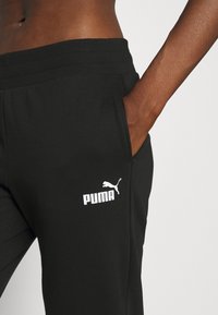 Pantalones de chándal negros con cintura elástica, que cuentan con un bolsillo lateral y un logo blanco de Puma en el muslo izquierdo. Textura de tela suave.