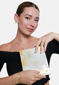 Goudaccentverpakking vastgehouden door een persoon in een zwarte off-shoulder top, gouden creolen en gestapelde armbanden, met zachte natuurlijke huidtinten.