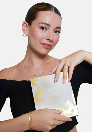 Une femme portant un top noir décolleté tient une boîte marbrée accentuée de doré, étiquetée "hedeman", mettant en avant un fini brillant et un design élégant.