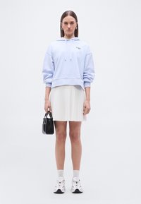 Femme portant un sweat à capuche bleu clair et une jupe plissée blanche, tenant un petit sac à main noir, debout devant un fond blanc uni.