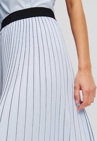 Jupe plissée bleu clair avec des rayures noires verticales, taille élastique noire avec logo, texture de tissu fluide. Main visible tenant la jupe.