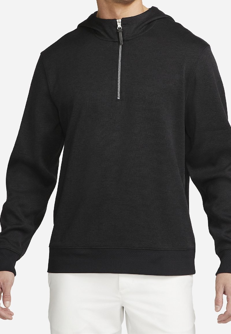 Homme portant un pull noir à manches longues avec une demi-fermeture éclair et une capuche, associé à un pantalon blanc, raccourci au niveau du cou et de la taille.