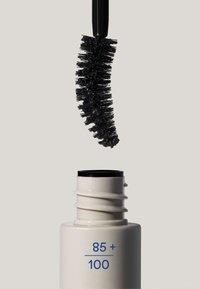 Una scovolino di mascara nero curvo sopra un tubetto beige aperto etichettato "85 + 100" su uno sfondo chiaro e neutro.