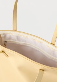 Sac fourre-tout jaune pâle avec deux poignées, montrant une poche intérieure zippée en mesh blanc et le nom de la marque "Lacoste" sur la bordure en tissu.