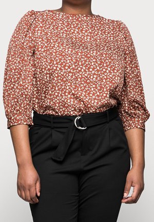 Femme portant un chemisier imprimé floral couleur rouille à manches bouffantes et un pantalon noir taille haute avec une ceinture en tissu et une boucle argentée.
