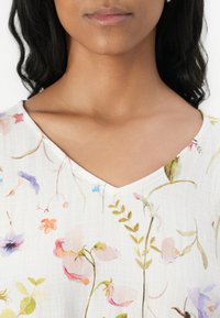 Lett hvid bluse med V-udskæring, prydet med et blomsterprint i bløde pasteller og delikate grønne accenter. Struktureret stof.