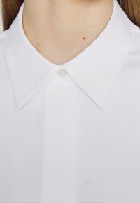 Acercamiento de una camisa blanca con cuello, abotonada en la parte superior, mostrando el área del cuello con dos pequeñas manchas en la piel.