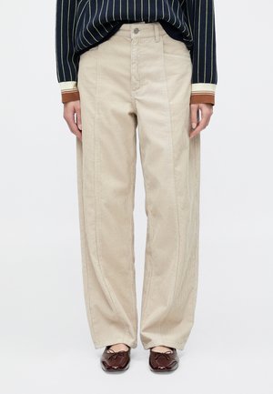 Beżowe szerokie spodnie z sztruksu z przednimi kieszeniami i widocznymi przeszyciami. Połączone z ciemną, paskowaną bluzką z długim rękawem i brązowymi butami.