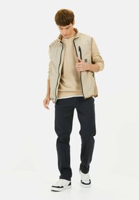 Lichtbeige ritsvest met een hoge kraag, gedragen over een lichte trui, gecombineerd met donkere broek en witte sneakers, met een logodetail op het vest.
