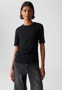 Donna con capelli corti e scuri, che indossa una maglietta nera aderente a maniche corte e jeans scuri a vita alta, in piedi contro uno sfondo chiaro e semplice.