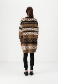 Cárdigan largo marrón con un patrón de diseños geométricos en negro, blanco y marrón; presenta un dobladillo acanalado y un corte holgado, combinado con leggings y zapatos negros.