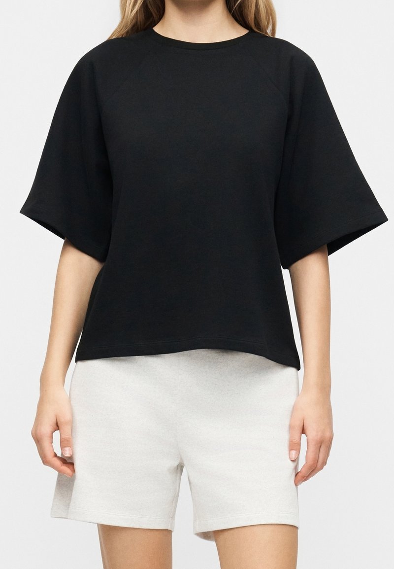 Femme portant un haut noir ample à manches courtes et un short décontracté gris clair sur un fond uni.