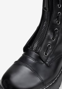 Botín de tobillo de imitación cuero negro con puntera redondeada, cordones frontales y cremallera lateral con herrajes plateados. Costuras blancas acentúan la suela.