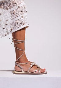 Sandalias planas doradas con un diseño de tiras, adornadas con perlas sintéticas, se muestran en un pie, con una delgada pulsera en el tobillo.