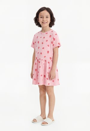 Barn står smilende, iført en rosa kjole med korte ermer og røde jordbærtrykk, kombinert med hvite slidesandaler.