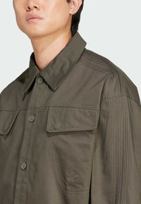 Giacca verde oliva con design a bottoni, ampie tasche sul petto e logo ricamato discreto. Dettagli a trama sulle spalle e tessuto liscio.