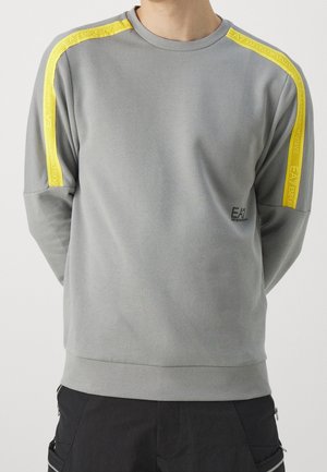 Mężczyzna noszący szarą bluzę z żółtymi paskami i logo "EA7 Emporio Armani" na ramionach i klatce piersiowej.