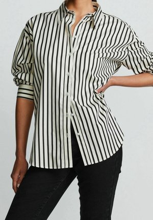 Personne portant une chemise blanche à rayures verticales noires et un pantalon noir, une main sur la hanche, sur un fond clair uni.