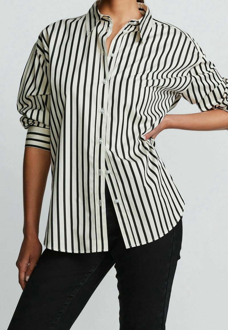 Personne portant une chemise blanche à rayures verticales noires et un pantalon noir, une main sur la hanche, sur un fond clair uni.