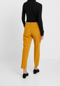 Żółte zwężające się spodnie o gładkiej fakturze, niskim stanie, z dwiema tylniami kieszeniami, zestawione z czarną bluzką z długim rękawem i czarnymi butami.