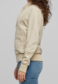 Femme portant une veste bomber beige clair avec des poignets élastiques et un jean bleu, vue de profil sur un fond uni.
