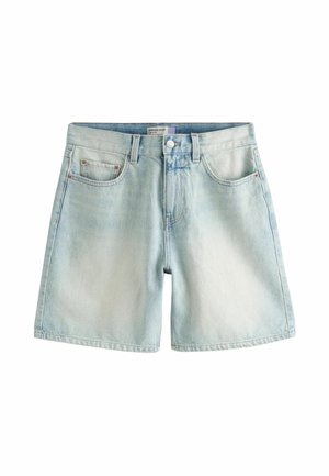 Person wearing light blue denim shorts and holding a white loose top above the waist against a neutral background.