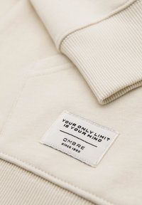 Poignet en tissu côtelé couleur crème près d'une étiquette cousue portant l'inscription « VOTRE SEULE LIMITE EST VOTRE ESPRIT » et « OMBRE Depuis 1985 » sur un sweat-shirt.