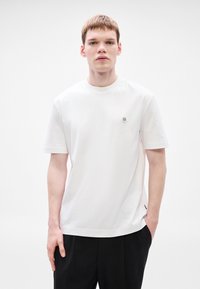 Biele bavlnené tričko s krátkymi rukávmi, okrúhlym výstrihom a malým kruhovým logom na ľavej strane na hrudi. Kombinované s čiernymi nohavicami.