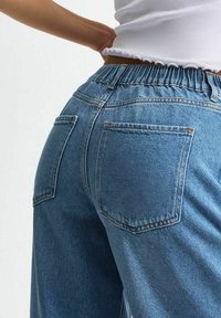 Primer plano de una persona que lleva jeans azules de cintura alta con cinturilla elástica y una blusa corta blanca con bordes con volantes.