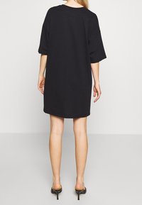 Czarna, oversize sukienka t-shirtowa, krótkie rękawy, okrągły dekolt, miękka bawełniana tekstura, lekko zaokrąglony dół, noszona z czarnymi obcasami mules.