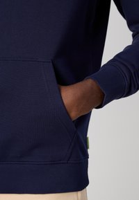 Sweatshirt bleu marine avec des poignets et un ourlet à côtes, doté d'une poche frontale. Le matériau semble doux et texturé, avec une étiquette sur le côté.