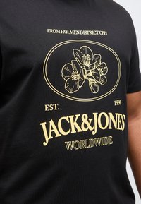 Μαύρο μπλουζάκι με κίτρινο λουλουδάτο σχέδιο και κείμενο που λέει "FROM HOLMEN DISTRICT CPH," "EST. 1990," "JACK&JONES," και "WORLDWIDE."