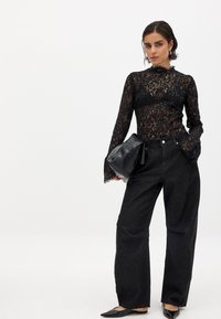 Schwarzspitzenshirt mit langen Ärmeln und Rüschenausschnitt, kombiniert mit hochtaillierten schwarzen Denim-Hosen mit weitem Bein. Schwarze Handtasche und spitze Ballerinas.