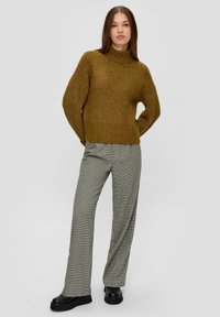Senfgelber Strickpullover mit hohem Kragen, weiten Ärmeln, kombiniert mit grauen Karohosen und schwarzen Plateauschuhen.