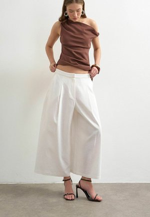 Frau passt weiße, weit geschnittene Hosen an, trägt ein braunes One-Shoulder-Top, schwarze Sandalen mit hohen Absätzen und auffällige Ohrringe vor einer schlichten Wand.