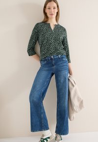 Femme portant un chemisier vert à motifs et un jean bleu à larges jambes, tenant une veste beige, debout devant un mur clair uni.