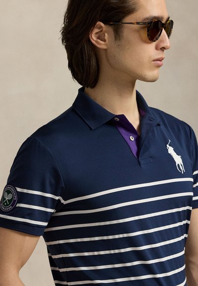 Férfi teknősbéka mintás napszemüvegben, tengerészkék csíkos galléros ingben, fehér logóval és Wimbledon-páccal az ujján.