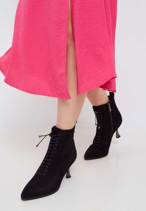 Stivali neri in suede alla caviglia con punte appuntite, lacci davanti e tacchi di 7 cm, indossati con un vestito rosa brillante e strutturato con uno spacco laterale.