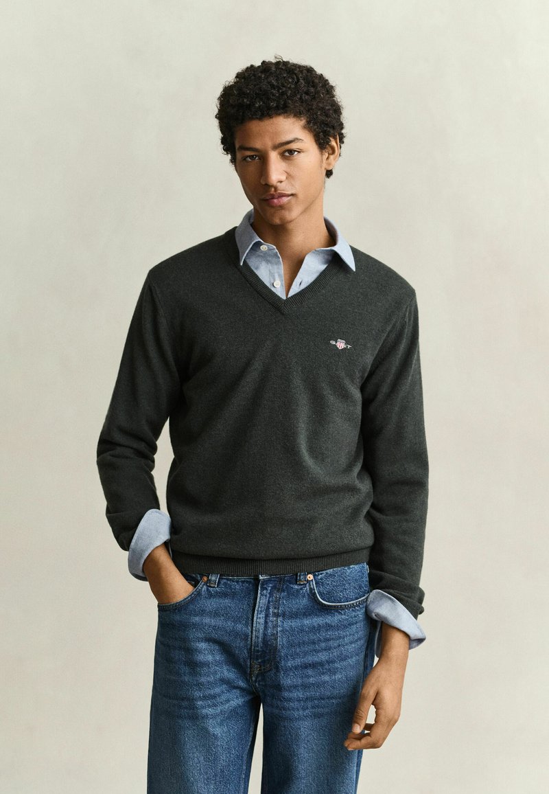 Jeune homme aux cheveux bouclés portant un pull à col en V foncé sur une chemise bleu clair et un jean bleu, debout avec une main dans la poche.