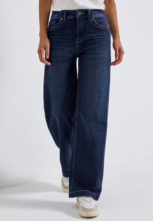 Brede donkere blauwe jeans van denim, met een hoge taille, steekzakken en zichtbare stiksels. Vergezeld van witte sneakers.