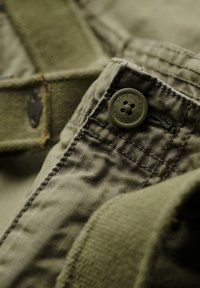 Olijfgroene stof close-up met een knop met "SUPERDRY" branding, getextureerde stiksels en duurzame riemdetails.