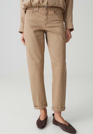 Frau trägt beige Jeans mit geradem Bein und umgeschlagenen Säumen, kombiniert mit dunkelbraunen spitzen Ballerinas vor neutralem Hintergrund.