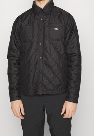 Homme portant une veste noire matelassée avec des boutons-pression et deux poches poitrine, dont une avec le logo The North Face.