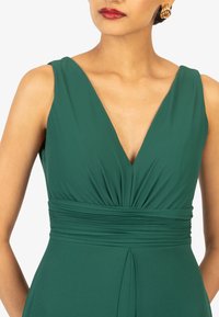 Grünes ärmelloses Kleid mit V-Ausschnitt, gerafftem Oberteil und strukturiertem Taillenbund. Glatter Stoff und dezente Drapierung im Rock.