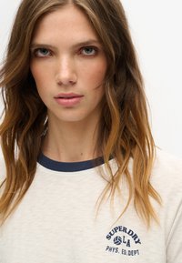 Cremefarvet kortærmet skjorte med marineblå ribbet halsudskæring, prydet med en blå "SUPER DRY LA" logo med en volleyball grafik. Blød tekstur.