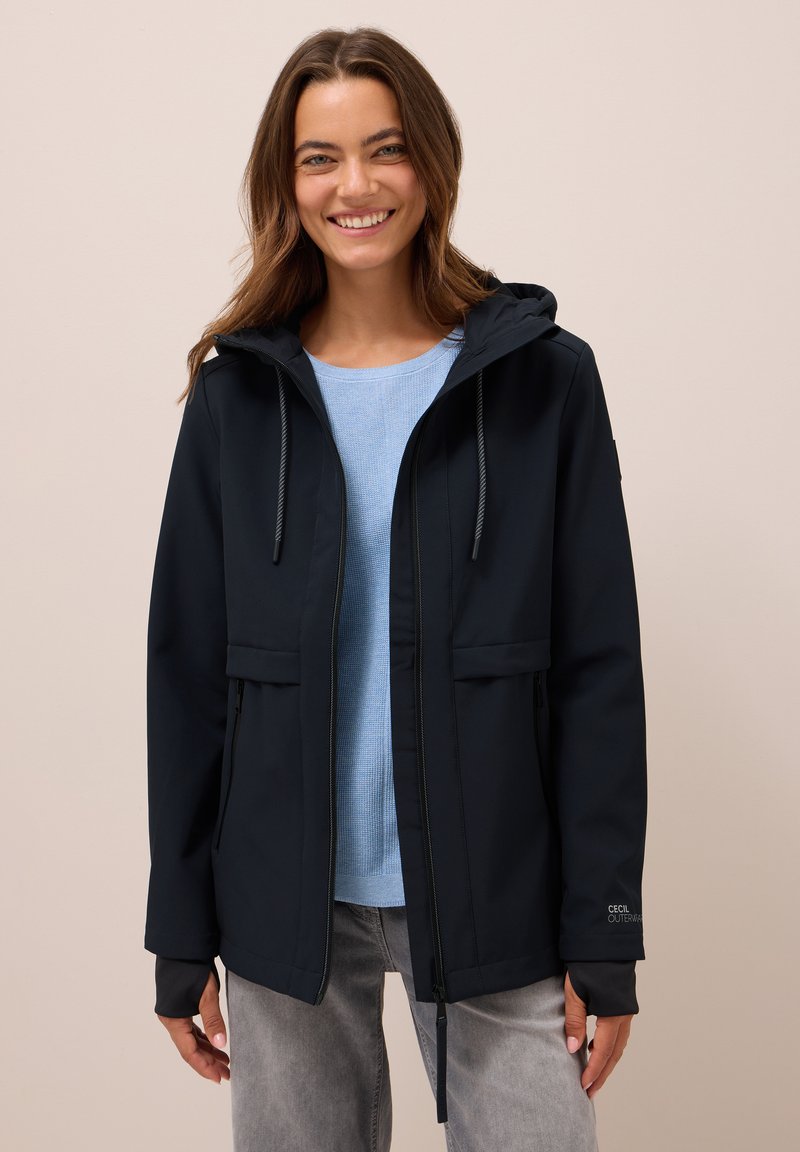 Marinblå softshell-jacka med huva, dragsko-stängning, två framfickor och elastiska muddar. Bärs över en ljusblå topp och grå jeans.